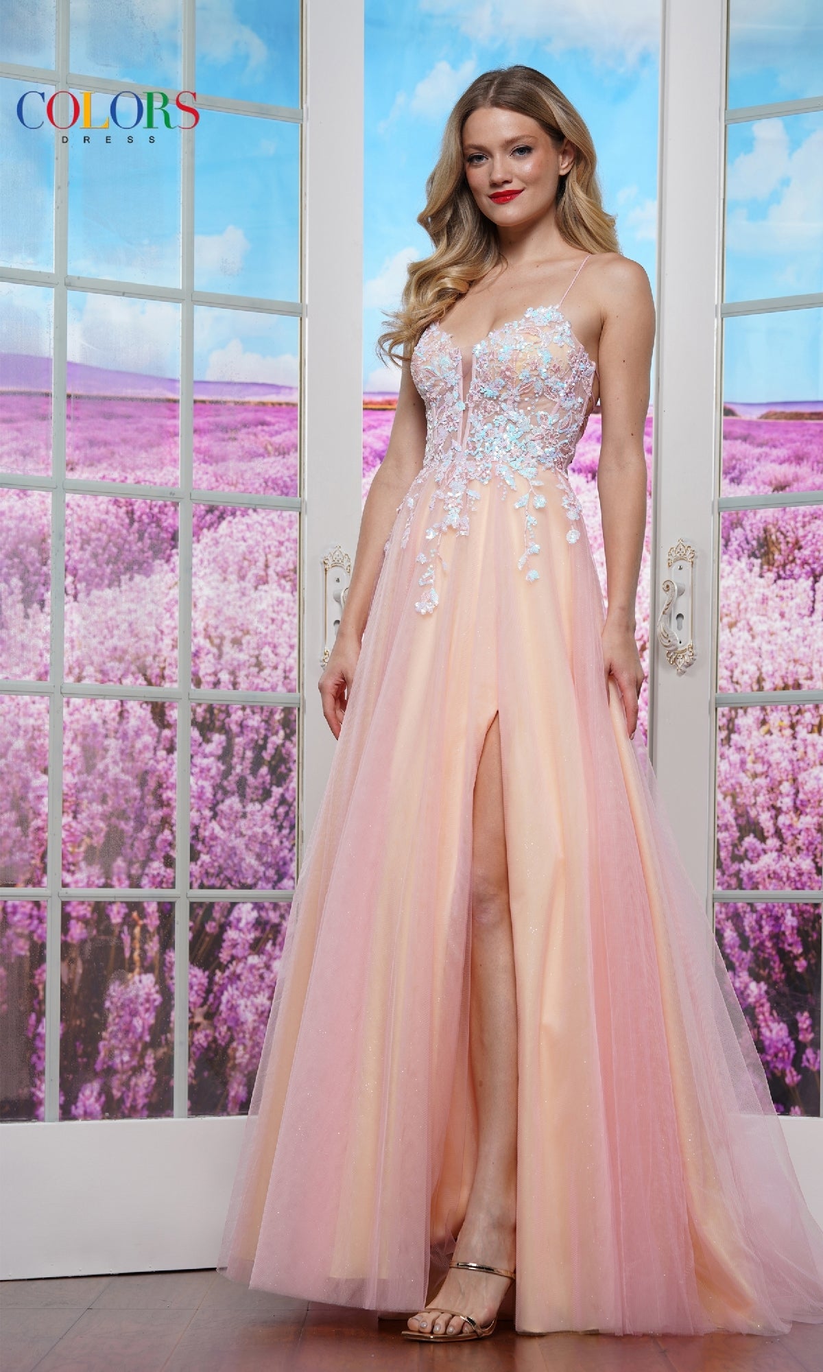 A woman wears the Strappy-Back Prom Ball Gown: Colors Dress 3497, a peach and blue floral A-line dress with a sheer bodice, standing indoors by glass doors overlooking a lavender field.